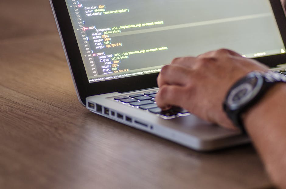 A close-up view of a person's hand operating a silver laptop on a wooden desk, with the screen displaying lines of code in a text editor. The person's wrist is adorned with a black watch, and their fi