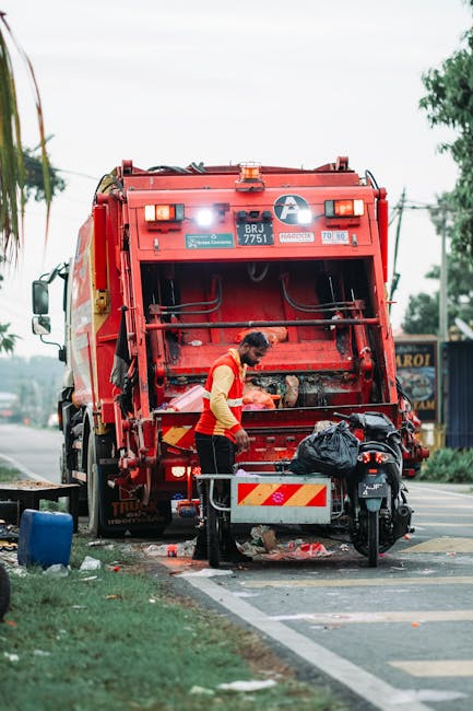 A waste collection worker wearing a high-visibility orange vest and black trousers is standing behind a large, bright red refuse collection truck on a roadside pavement during the daytime. The back of