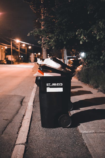 A waste collection worker wearing a high-visibility orange vest and black trousers is standing behind a large, bright red refuse collection truck on a roadside pavement during the daytime. The back of