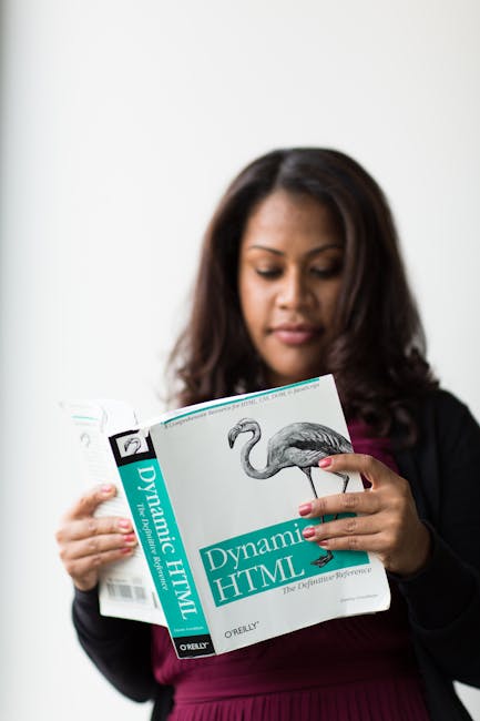 A woman with dark, shoulder-length hair and medium skin tone is seen holding and reading a book titled 'Dynamic HTML' with a flamingo illustration on the cover. She is dressed in a black blazer over a