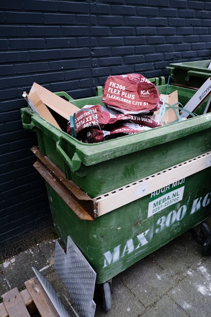 A black, cylindrical public waste bin made of metal with a domed lid and a slot for waste collection, situated on a cobblestone street. The surface of the bin appears weathered, with visible signs of